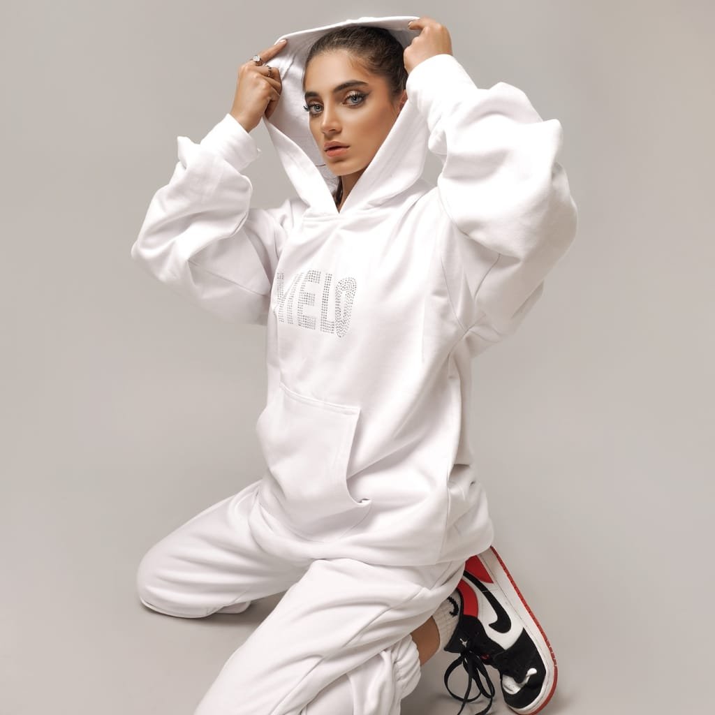 A young woman with styled hair and makeup is kneeling on the floor against a neutral background, wearing a white oversized hoodie and matching sweatpants, paired with black and white athletic shoes with red accents.