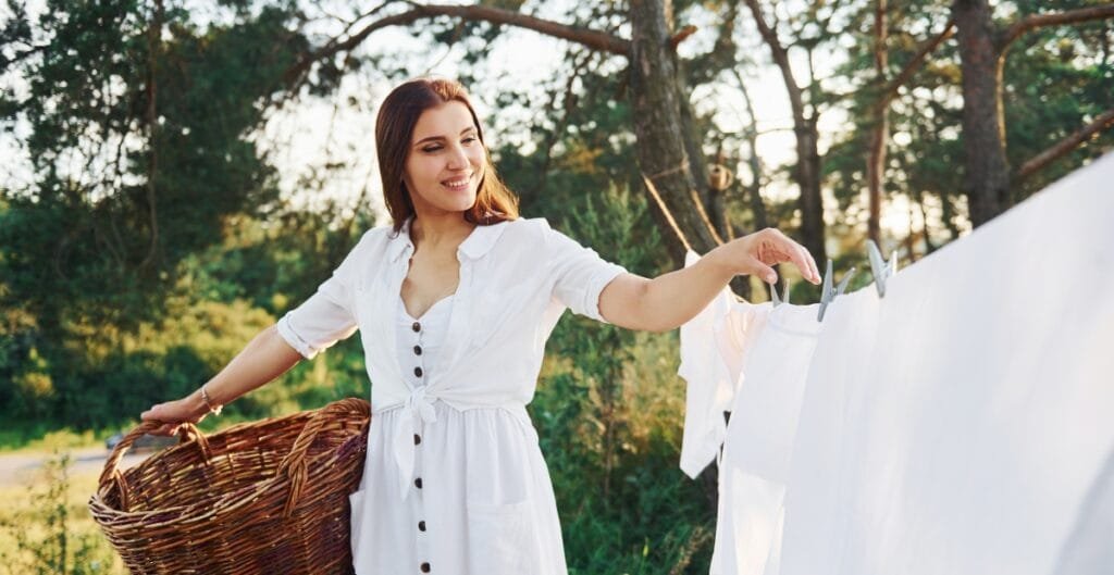 A woman in a white dress hanging laundry on a clothesline outdoors, holding a basket with one hand and using the other to clip clothes on the line, with trees and sunlight in the background.