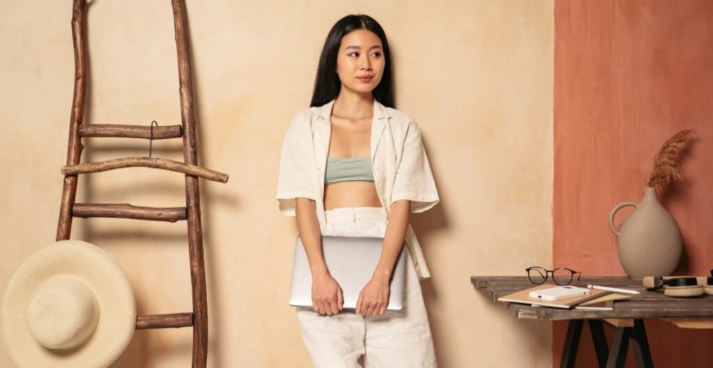 A young woman with long black hair, wearing a light green crop top and white outfit, stands against a textured beige and terracotta wall, holding a silver laptop. She is positioned near a rustic wooden ladder, a wide-brimmed straw hat, and a table with eyeglasses, a notebook, and a vase with dried grass.