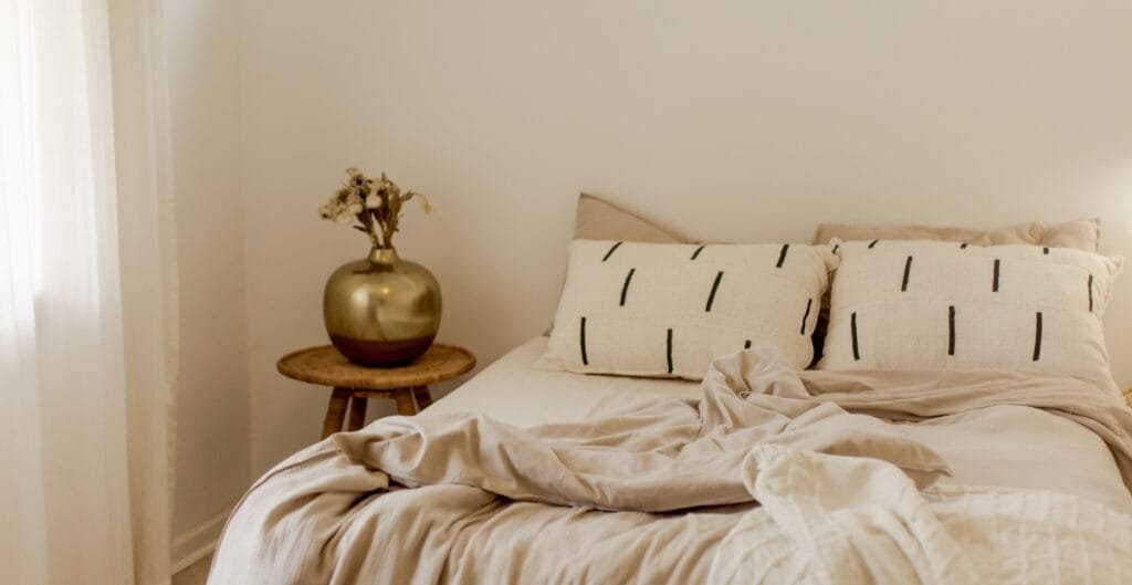 A neatly made bed with beige and cream-colored bedding, two pillows with black line patterns, and a small wooden side table holding a gold vase with dried flowers in a softly lit room.