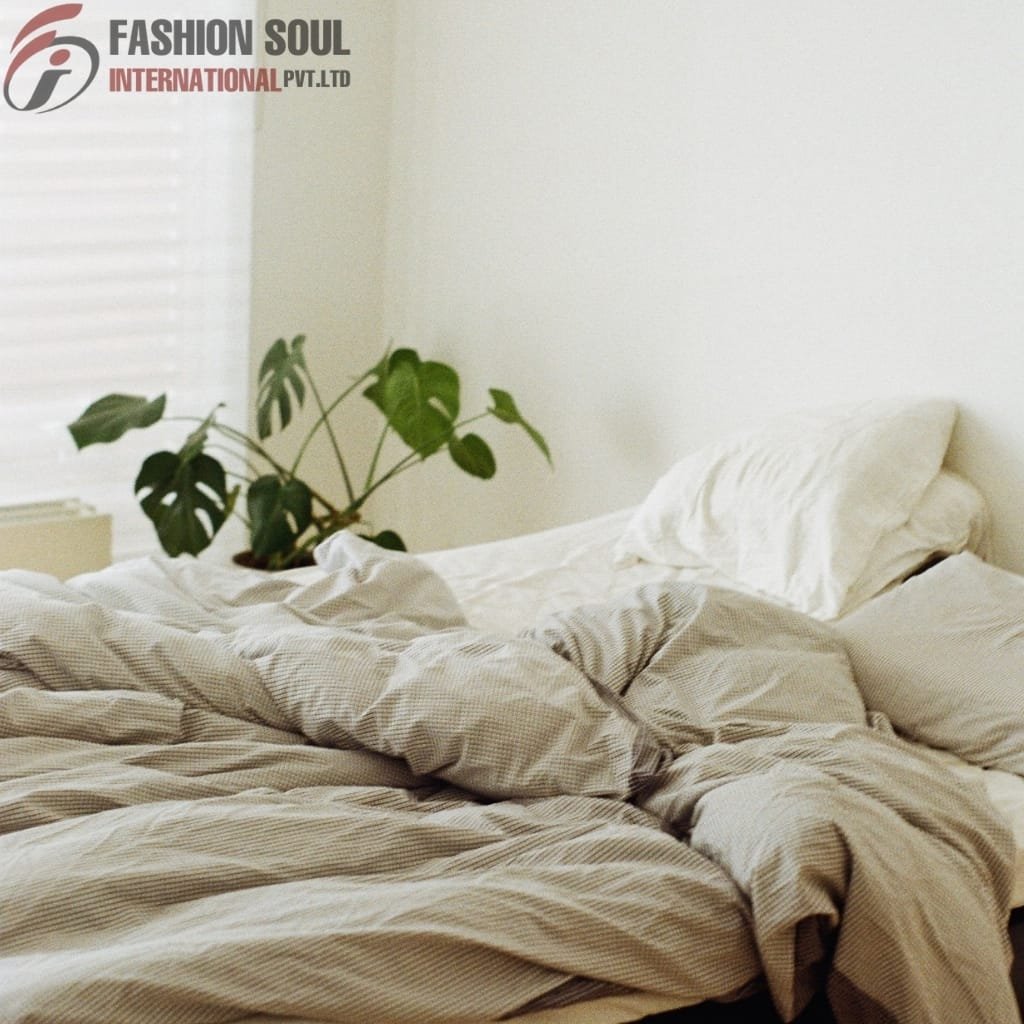 A cozy unmade bed with beige striped sheets, a white pillow, and a green houseplant on a white bedspread, in a bright room with a white background and blinds.