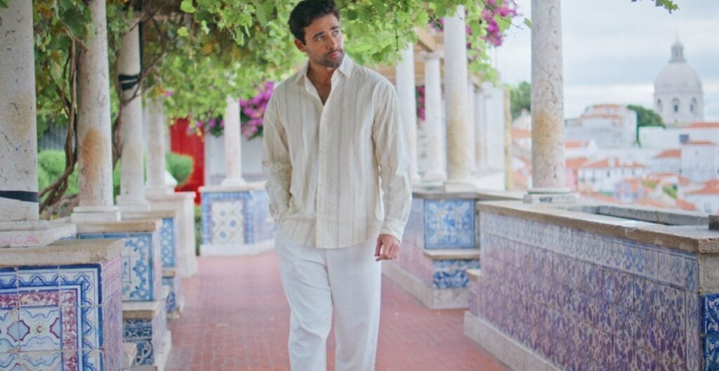 A man dressed in light-colored, relaxed clothing walking along a decorative outdoor colonnade with colorful tiles and greenery, overlooking a scenic view of a town with domed buildings.