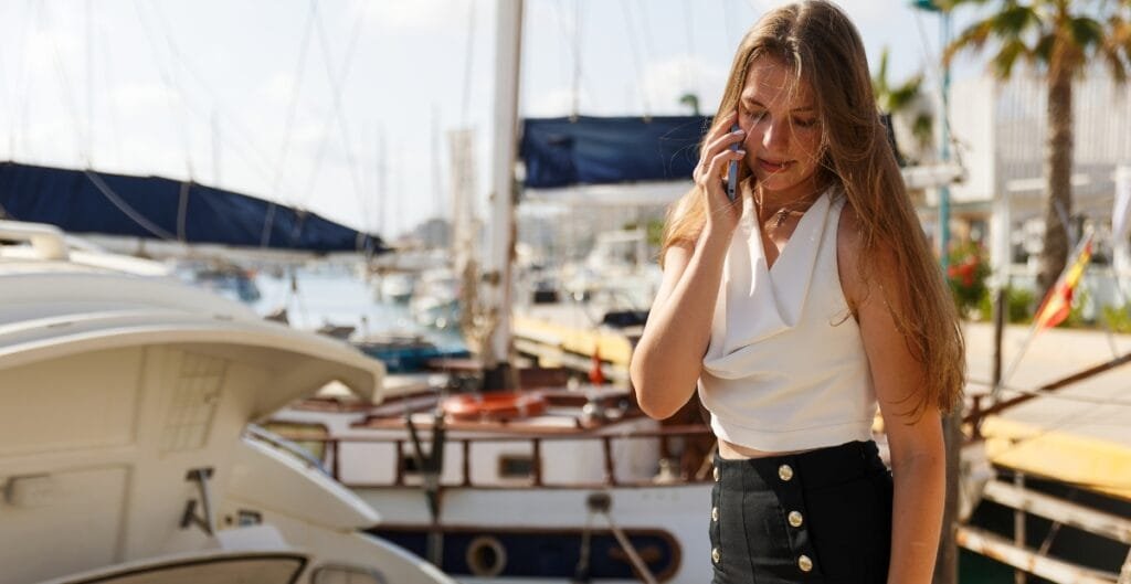 A young woman with long hair talking on her phone at a marina, surrounded by boats and yachts on a sunny day.