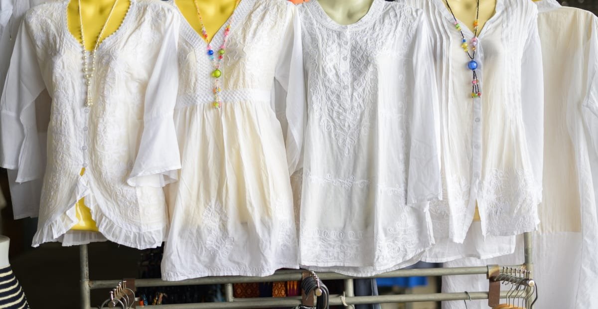A row of white embroidered blouses hanging on a clothing rack, with some featuring colorful necklaces, set against a yellow background.