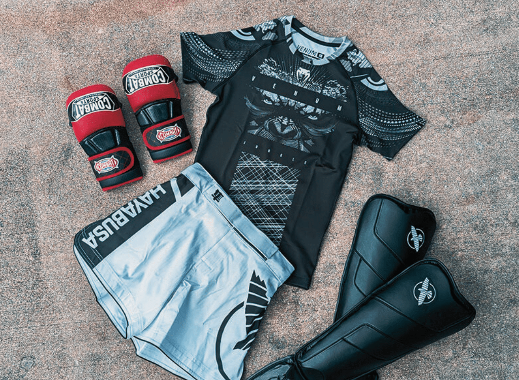 A collection of mixed martial arts (MMA) gear including gloves, shorts, a fight shirt, and shin guards laid out on a textured surface.
