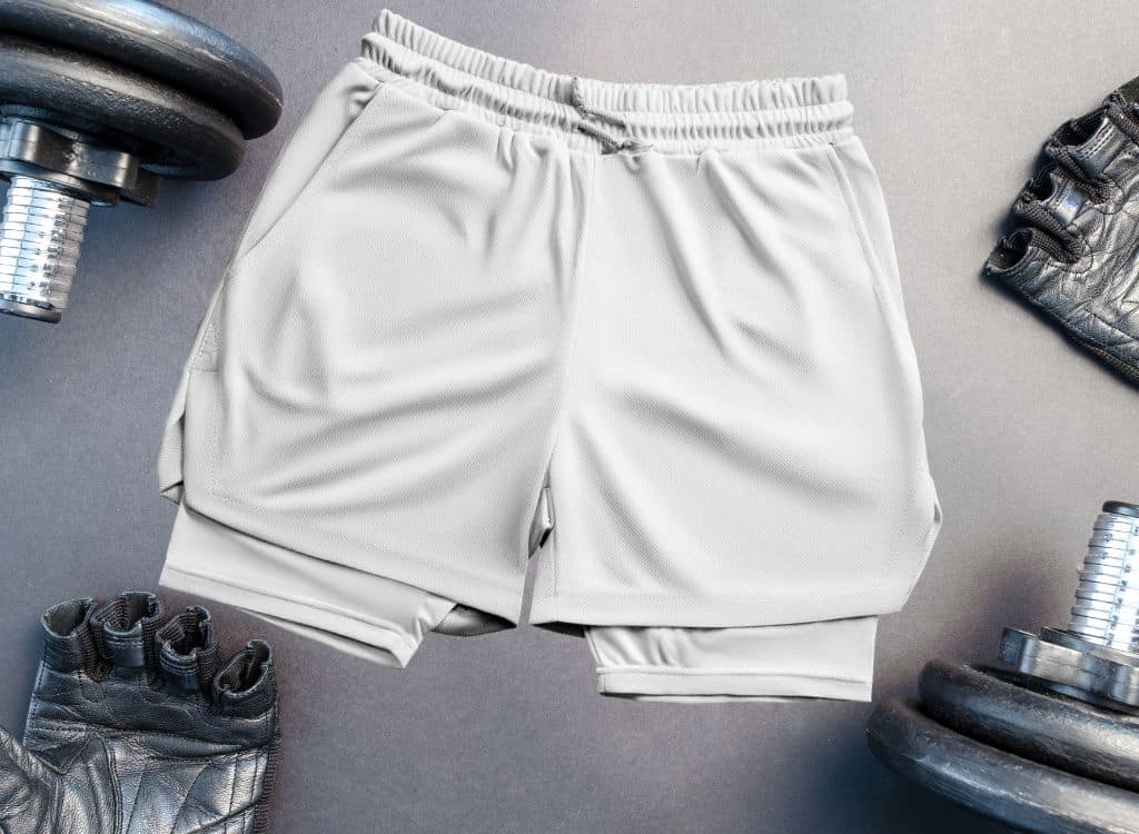 A pair of light gray MMA shorts laid flat on a gym surface, surrounded by dumbbells and workout gloves.
