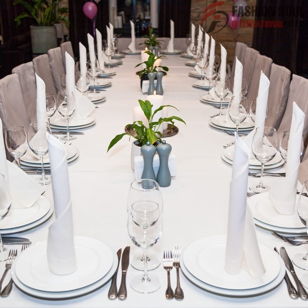 A long rectangular dining table set for a formal event, covered with a white tablecloth. The table is decorated with potted green plants and candles, with each place setting including a white plate, a rolled white napkin, and glassware. Beige upholstered chairs surround the table.