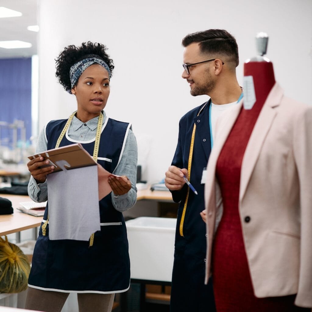 Three individuals are engaged in a conversation in a professional setting, with one woman holding a fabric sample and a clipboard, and two men listening and taking notes. A mannequin wearing a beige blazer and red top is visible in the foreground on the right.
