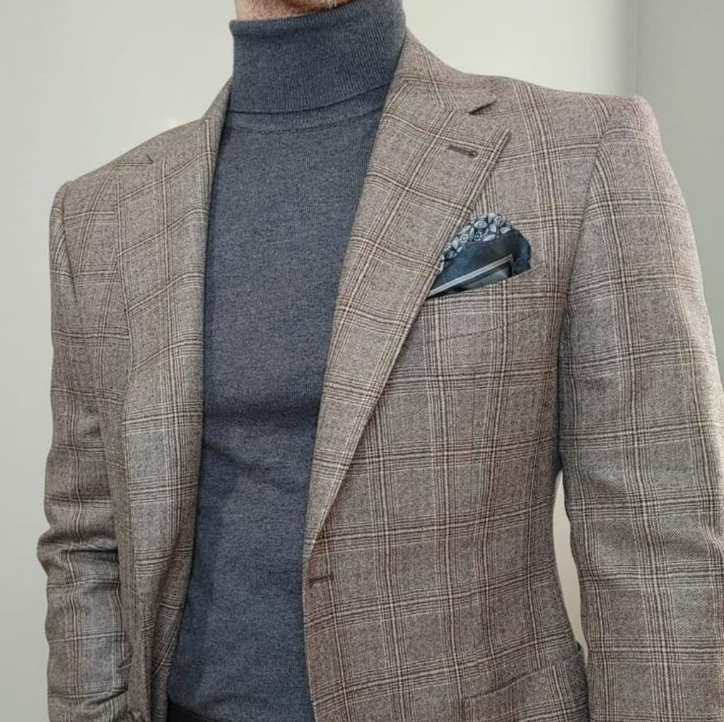 A man wearing a dark gray mock neck shirt underneath a brown plaid sport coat, with a patterned pocket square in his breast pocket.