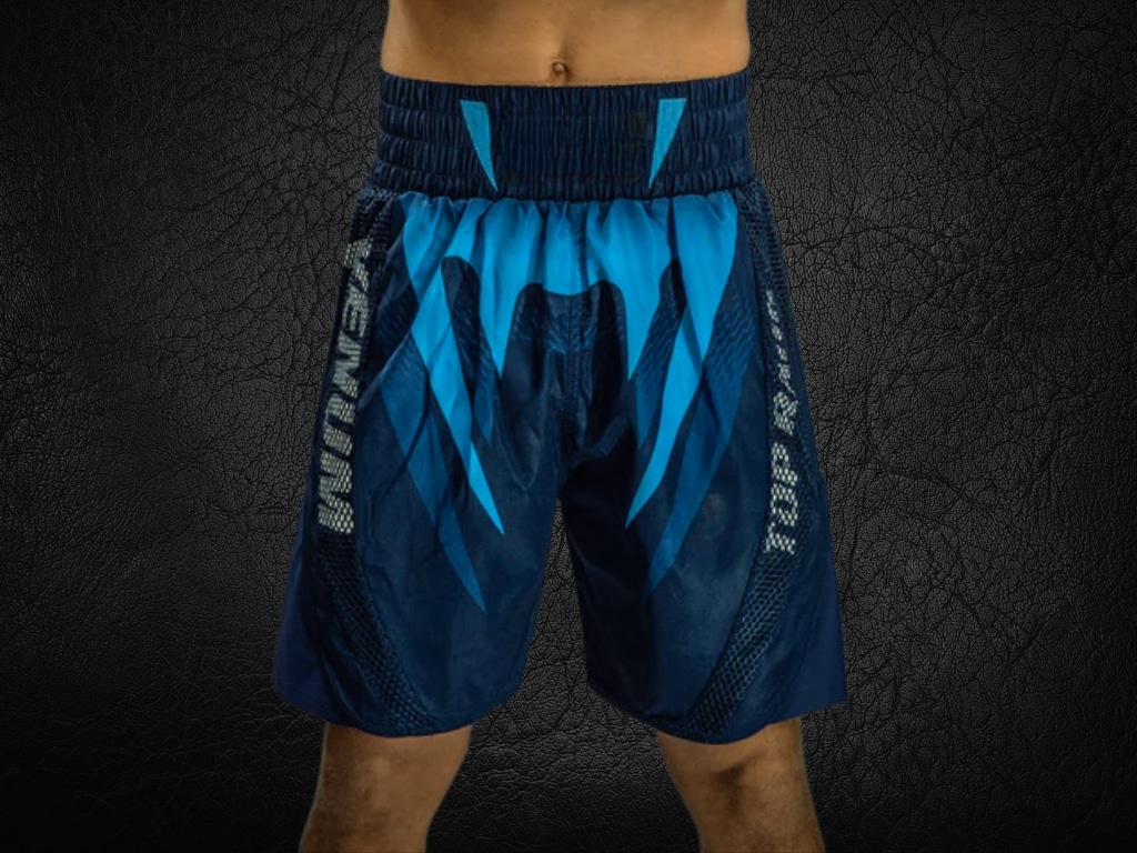 A close-up photo of blue boxing shorts with graphic design and branding, worn by a person standing against a dark textured background.