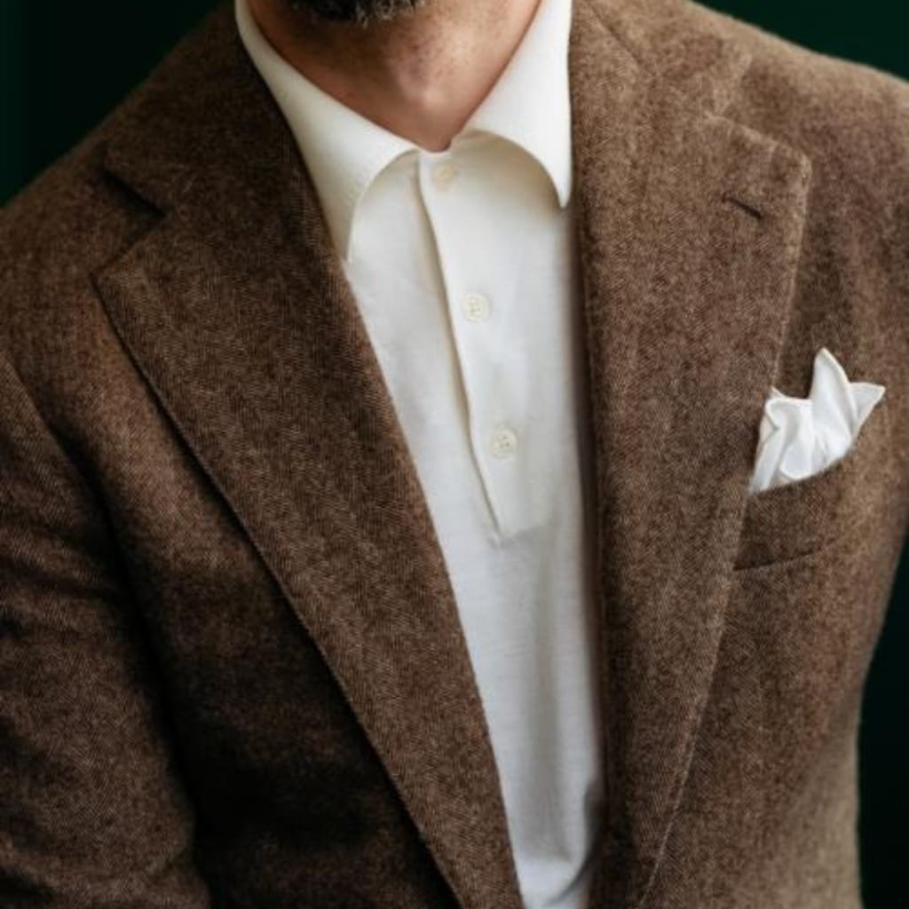 Close-up of a man dressed in a brown tweed blazer, white dress shirt, with a white pocket square. Only the upper part of his face and shoulders are visible, set against a dark background.