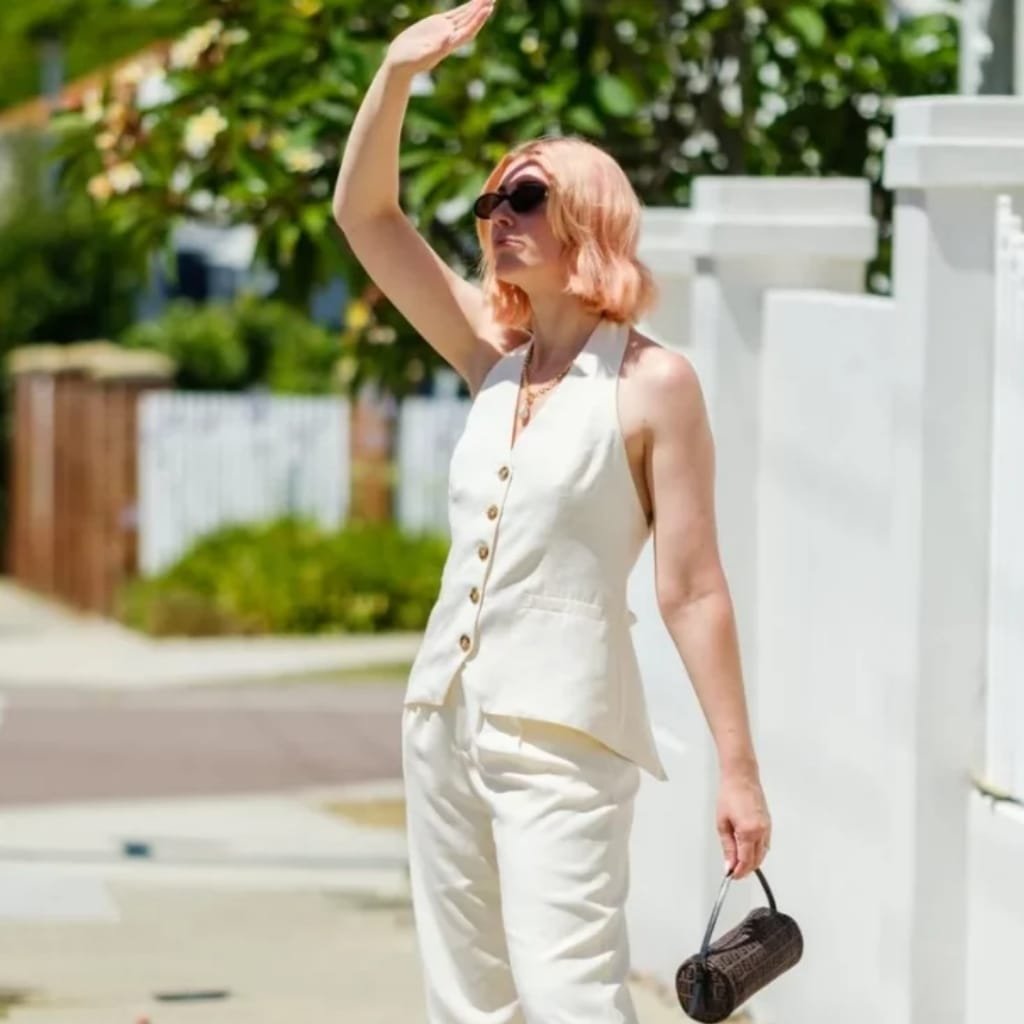  A woman with light hair and wearing sunglasses, dressed in a sleeveless cream-colored top and matching pants, standing outdoors on a sunny day. She is holding a small cylindrical bag in one hand and shielding her eyes with her other hand, as she looks into the distance. Green foliage and a white fence are visible in the background.