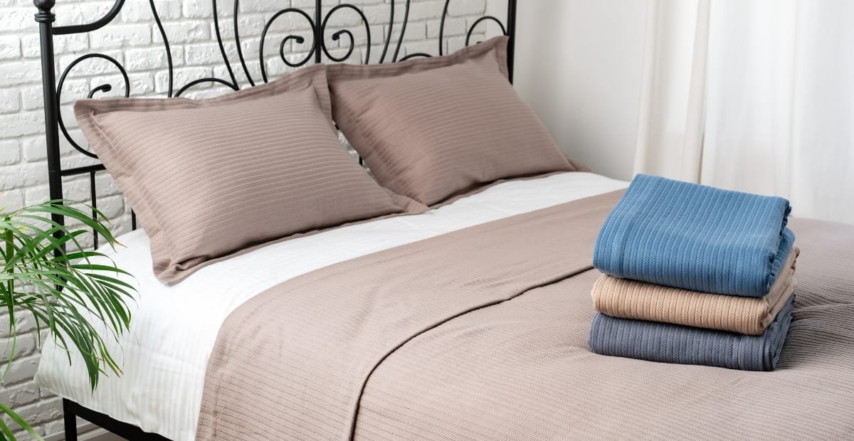 A neatly made bed with beige and brown bedding, two matching pillows, a small green plant on the left, and a stack of folded sweaters in blue, beige, and gray on the right.