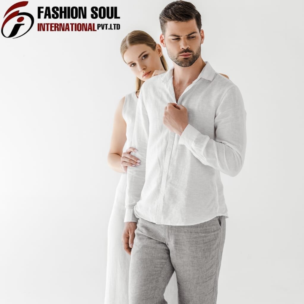 A man and woman dressed in light-colored, casual linen outfits standing against a plain white background. The man is in front, looking thoughtful with his hand near his chest, and the woman is slightly behind him, gently resting her hand on his waist while looking at the camera.