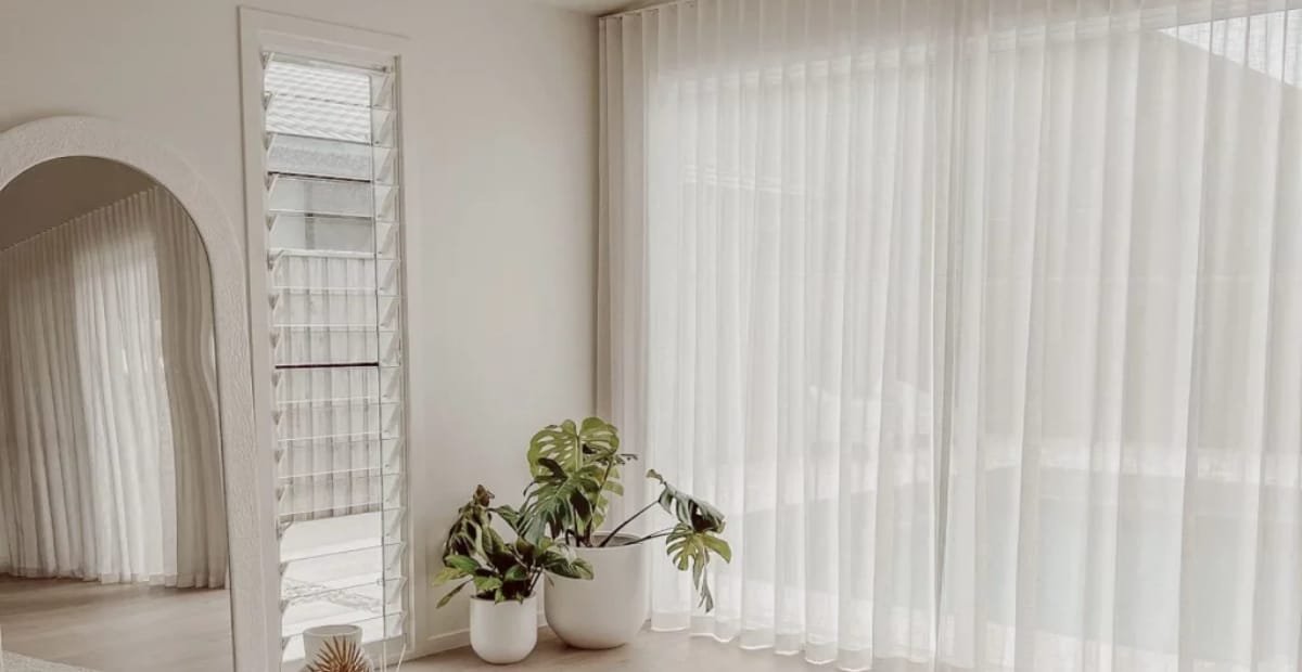 Bright interior corner with sheer white curtains, a large window, and three potted houseplants near the window.