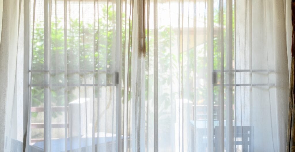 A view through sheer white curtains gazing out onto a patio with a table and chairs surrounded by greenery.