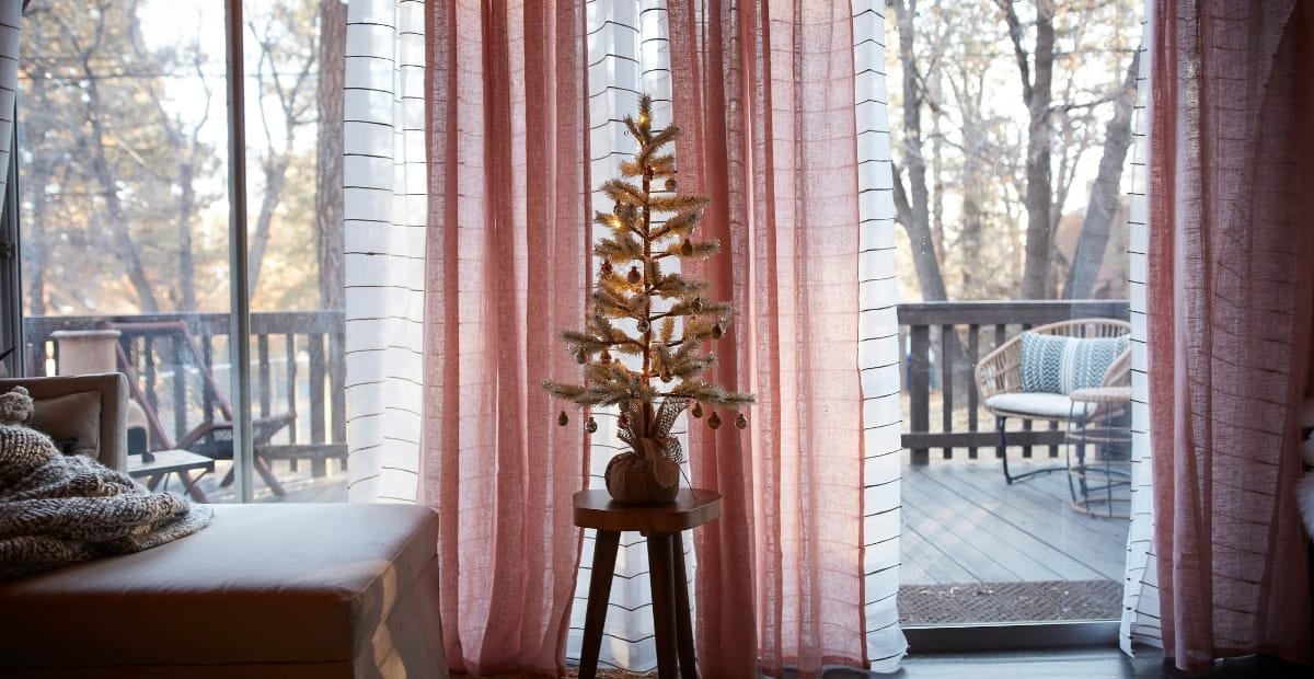 A cozy interior scene featuring a small decorated Christmas tree on a stand, placed in front of pink and white curtains that open to a wooden deck with outdoor furniture and trees in the background.