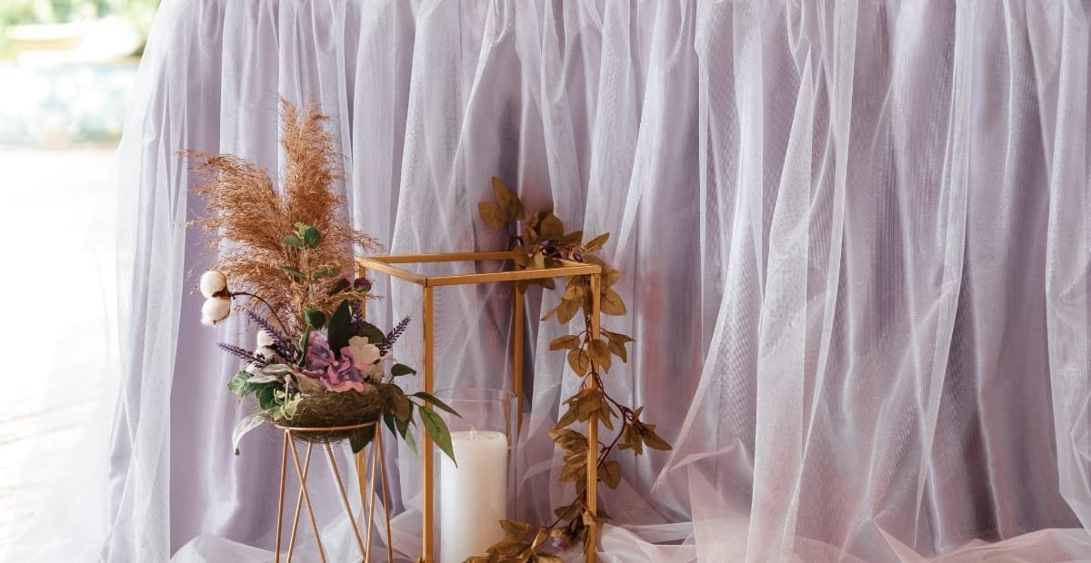 A floral arrangement with dried and fresh flowers displayed in a gold geometric vase, placed next to a white candle, against a backdrop of soft, sheer pink fabric.