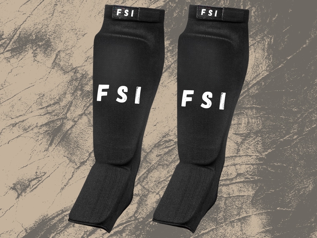 Pair of black MMA shin guards with "FSI" branding, placed on a textured surface.