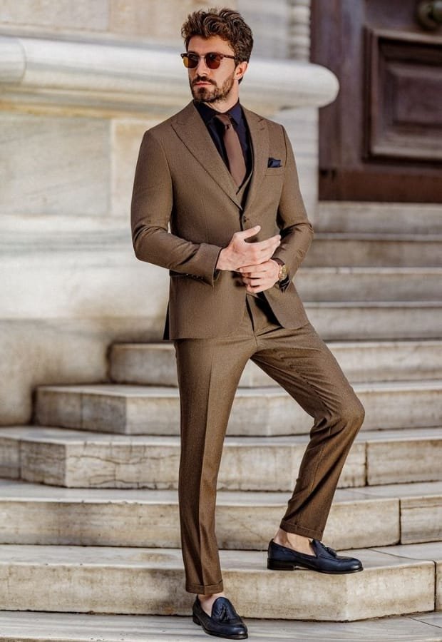 A man wearing a stylish brown suit with a matching sport coat, black shirt, and tie, standing on stone steps outdoors. He is holding his smartphone and wearing sunglasses.