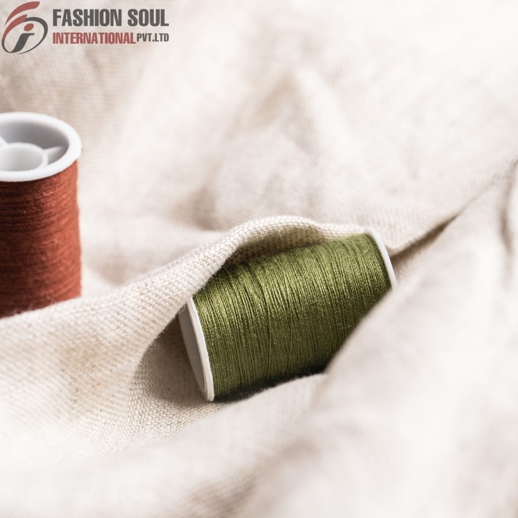 Two spools of thread, one green and one reddish-brown, resting on cream-colored fabric with a textured surface. The top left corner features the logo and name "Fashion Soul International Pvt. Ltd."