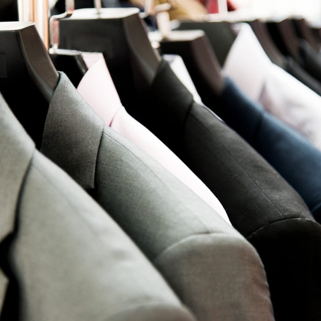  A row of hanging suits on black hangers, in various shades of gray, black, and a hint of pink, displayed closely together in a clothing store.