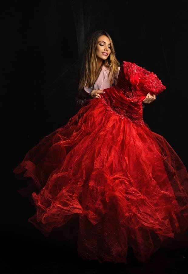 A woman in a stylish beige blazer holding and admiring a stunning red ball gown with intricate beading and voluminous tulle layers, against a dark background.