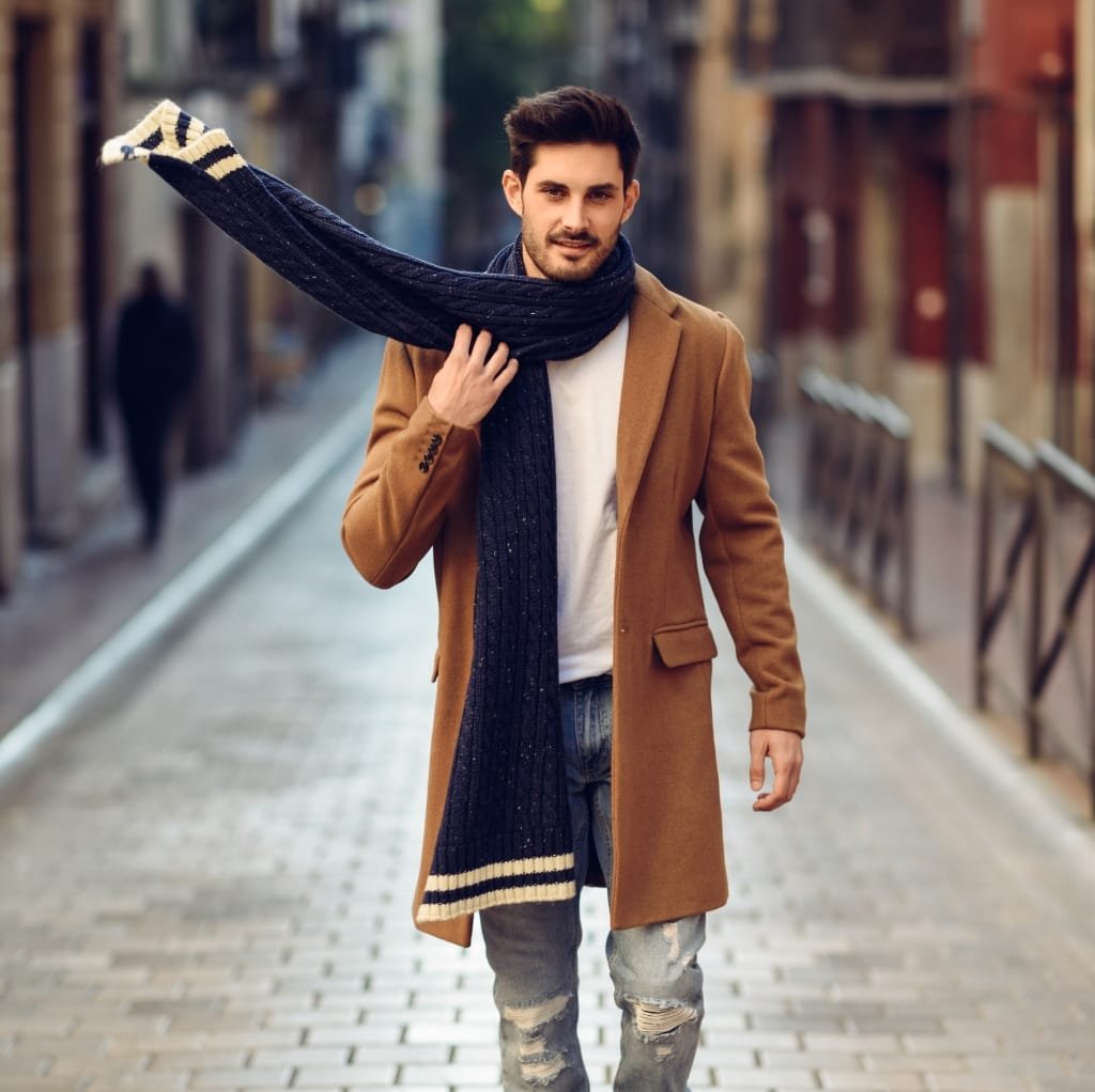 A stylish man walking down a city street wearing a tan sport coat, distressed jeans, and a dark navy scarf draped around his neck and over his shoulder.