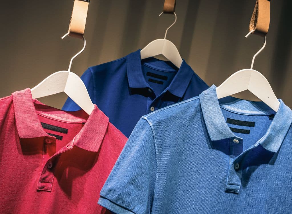 Three polo shirts in red, navy blue, and light blue with white labels hanging on display.