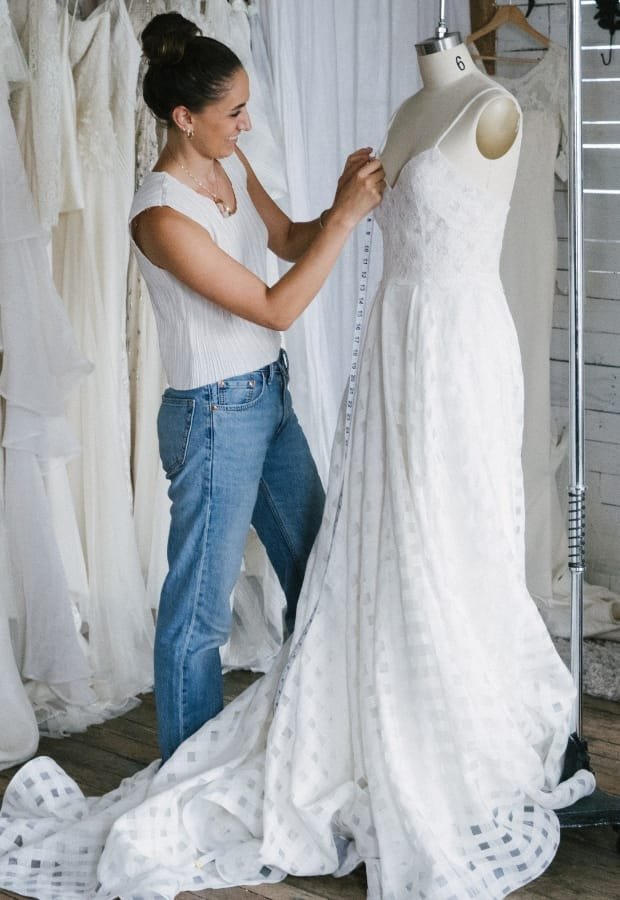 A woman measuring a white ball gown on a mannequin, inside a workshop or boutique with fabric swatches hanging in the background.