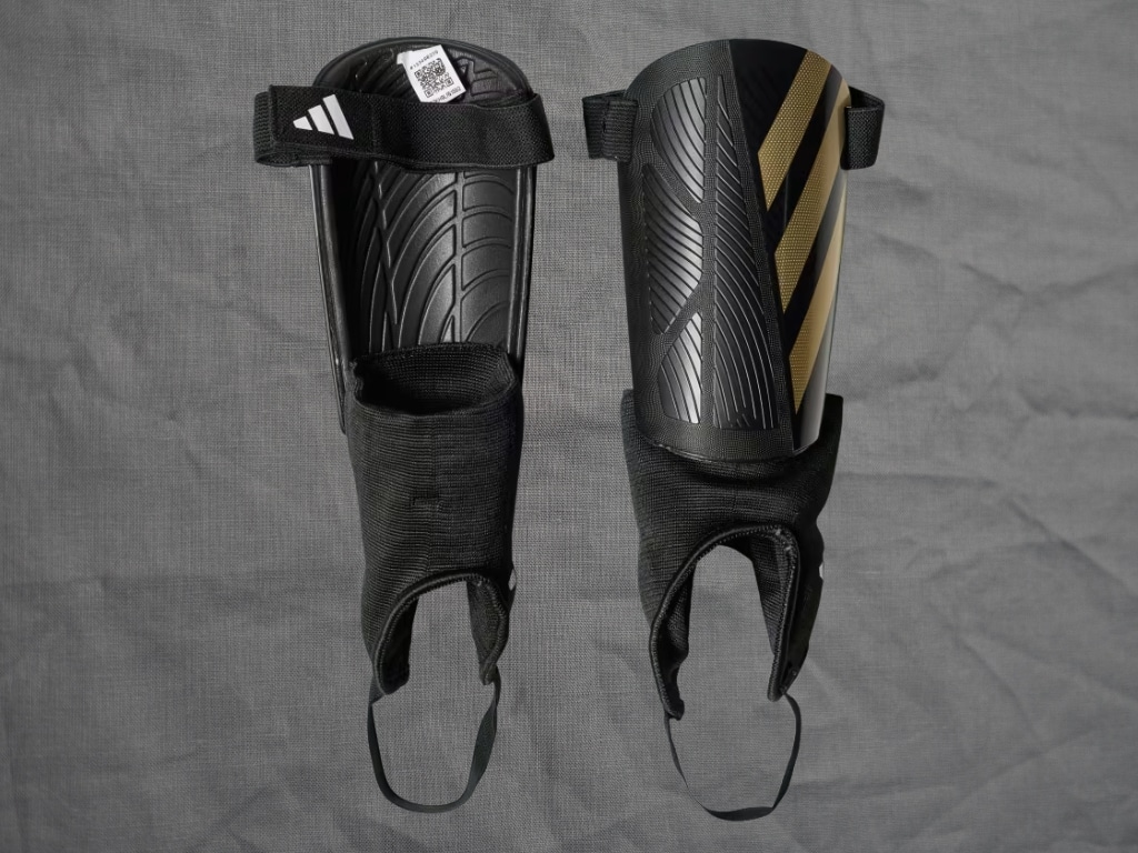 Pair of black MMA shin guards with textured padding and adjustable straps, placed on a gray fabric background.
