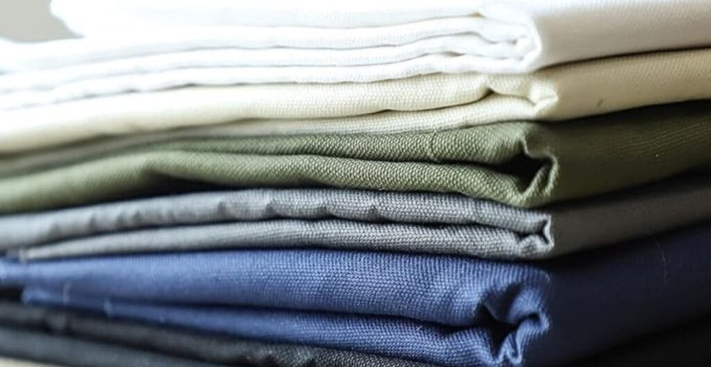 A stack of neatly folded duck cotton fabric in various solid colors, including white, cream, green, grey, blue, and black.