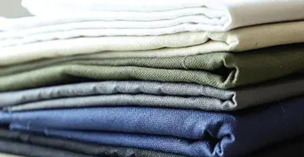 Stack of neatly folded fabric in various solid colors, including white, beige, green, grey, and navy blue.