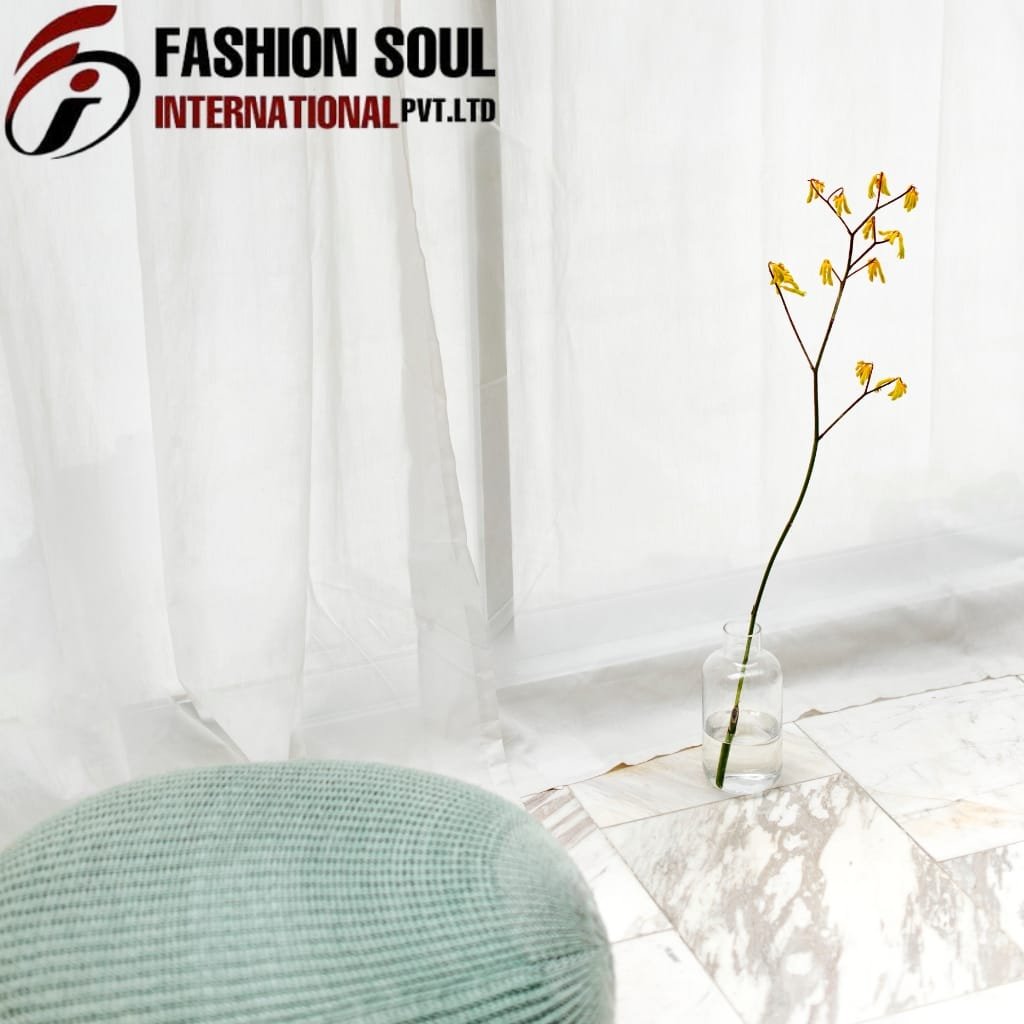  A minimalist scene featuring a single yellow flower with long stems in a clear glass vase placed on white marble tiles, against a backdrop of white curtains, with a portion of a light-colored, textured round cushion visible in the foreground.