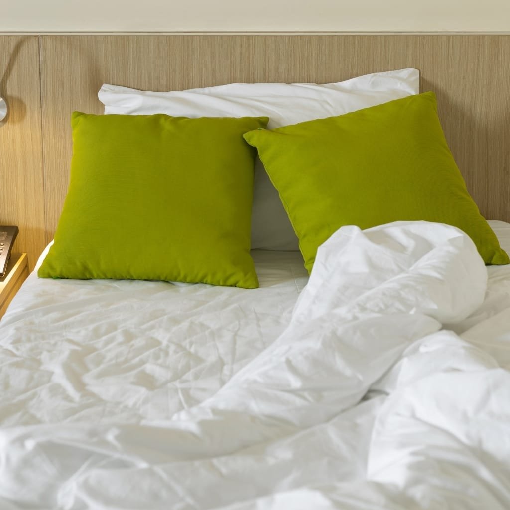 A neatly made bed with white sheets, two green pillows, and a white duvet. The headboard is wooden, and part of a bedside table with a telephone is visible.