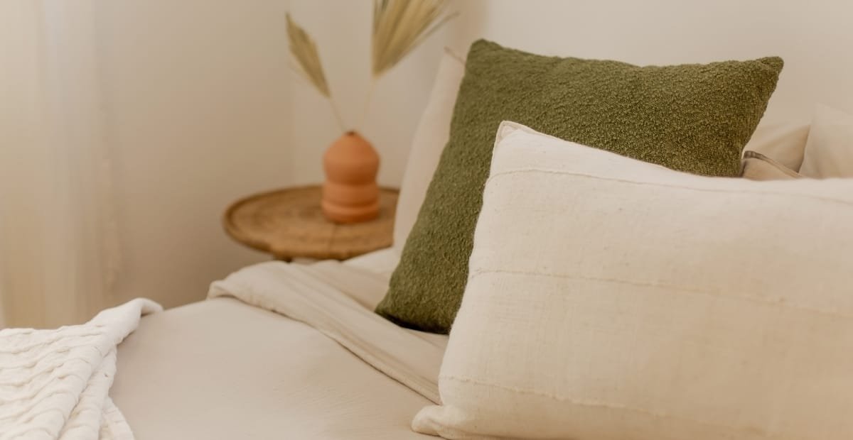 A cozy bedroom corner featuring a neatly made bed with white and cream pillows, one prominent green textured pillow, and a soft white blanket. In the background, there is a round wooden side table with a small, minimalist vase holding dried grasses, against a neutral wall.