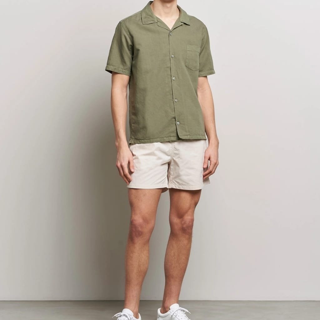A person wearing an olive green short-sleeve button-up shirt, beige shorts, and white sneakers standing against a plain light gray wall.