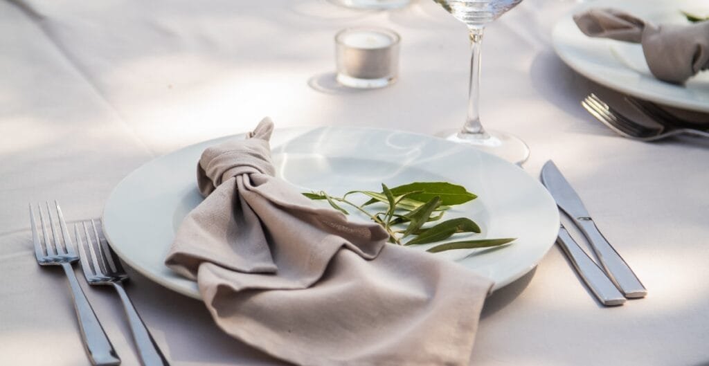 A elegantly set dining table with a white plate, beige cloth napkin decorated with green leaves, silver cutlery, a wine glass, and a small candle in the background.