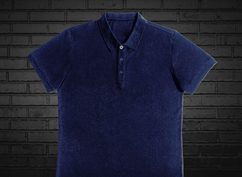 A plain white polo shirt with a small label on the chest, displayed against a dark dark brick background.