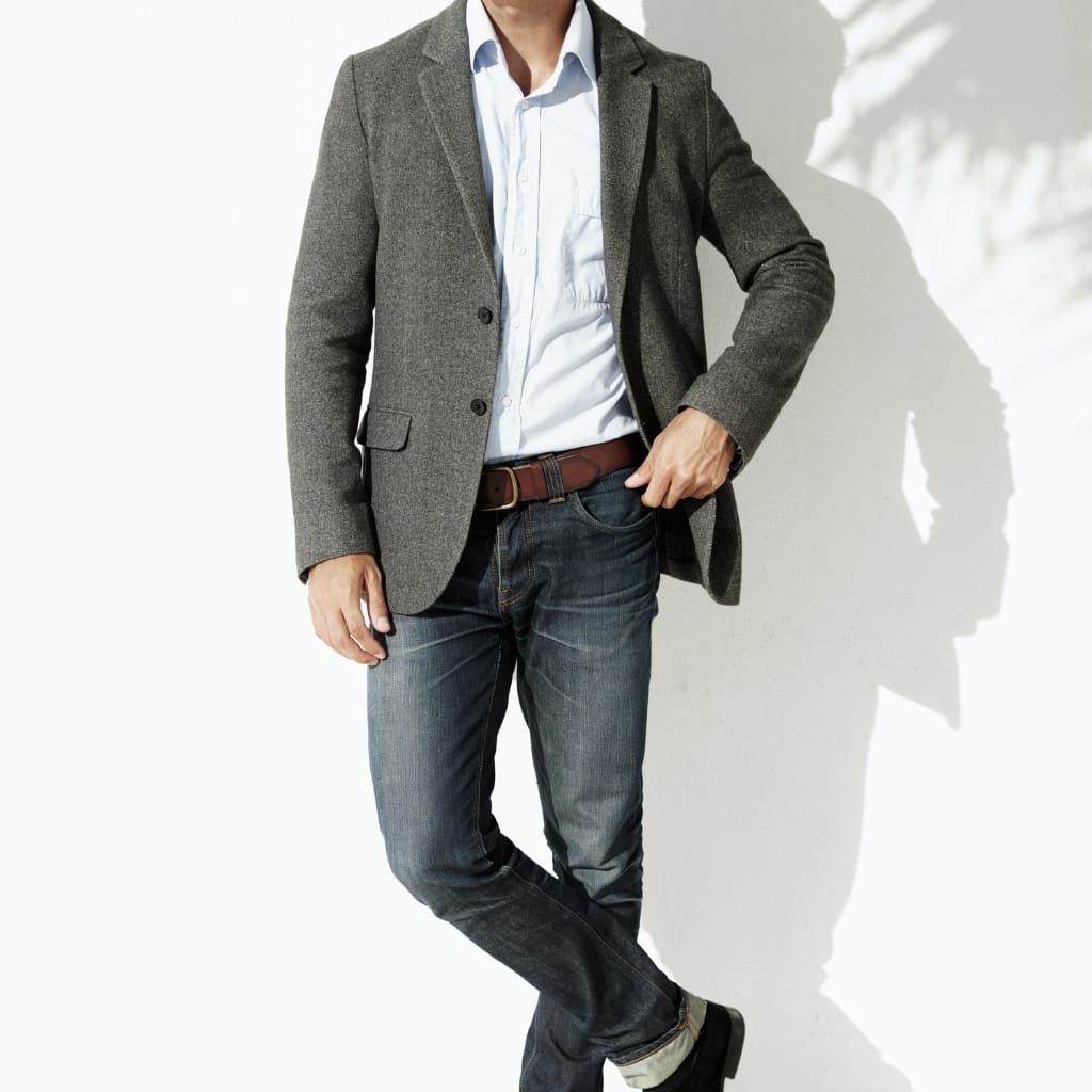 A man dressed in a smart-casual outfit wearing a grey sport coat over a light blue shirt, paired with dark jeans and black shoes, standing against a white background with shadows casting on the wall.