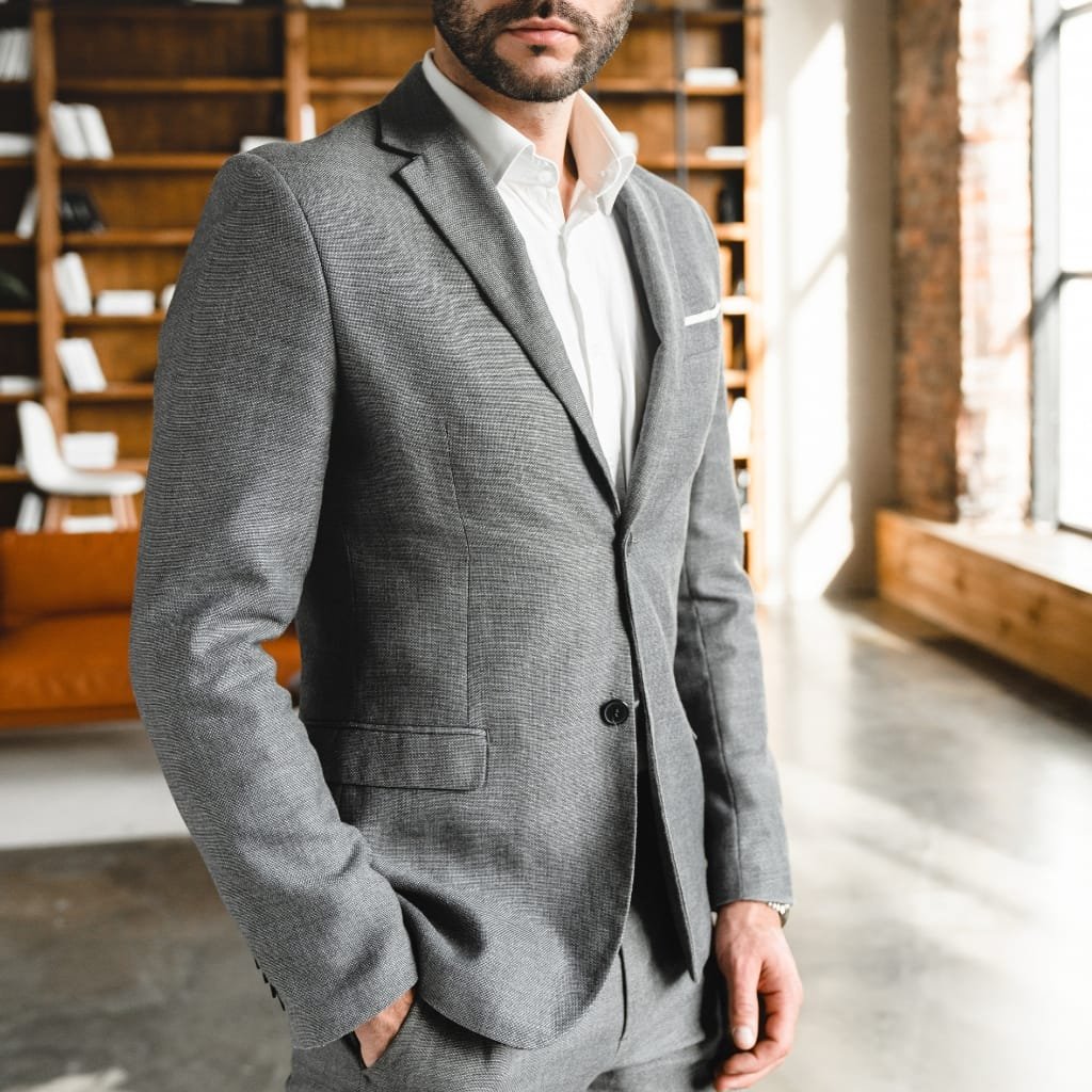 A well-dressed man wearing a grey sport coat and white shirt standing indoors, with a background featuring wooden shelves and a large window.