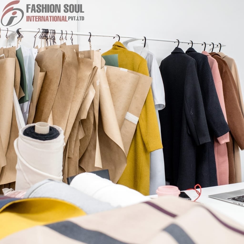 Clothing workspace with colorful fabric and finished garments hanging on a rack, including a yellow coat, black coat, and brown coat, with spools of fabric and related materials in the foreground.