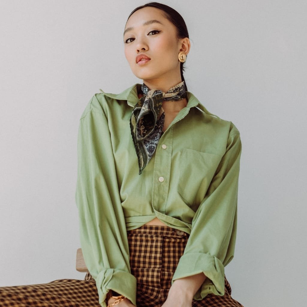 A stylish young woman with dark hair pulled back, wearing a light green oversized button-up shirt, a patterned scarf tied around her neck, gold earrings, and checkered brown and black pants, sitting against a plain light background with a confident expression.