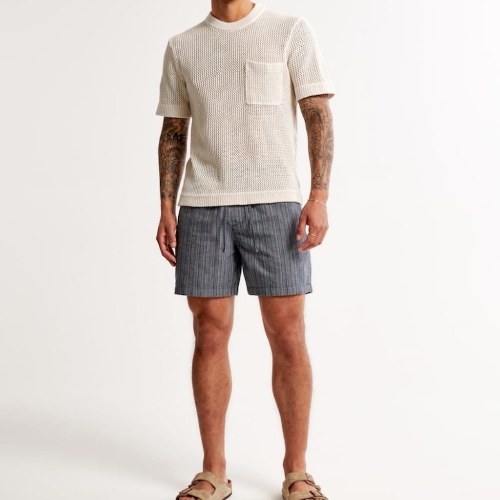 A person wearing a cream-colored knitted short-sleeve top with a pocket on the chest, paired with loose-fitting gray striped shorts and beige sandals, standing against a plain white background.