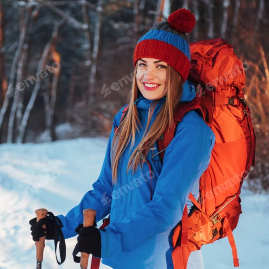 A smiling woman dressed in winter clothing, including a red, blue, and gray knit hat, a blue jacket, black gloves, and carrying an orange backpack, standing outdoors in a snowy forest.