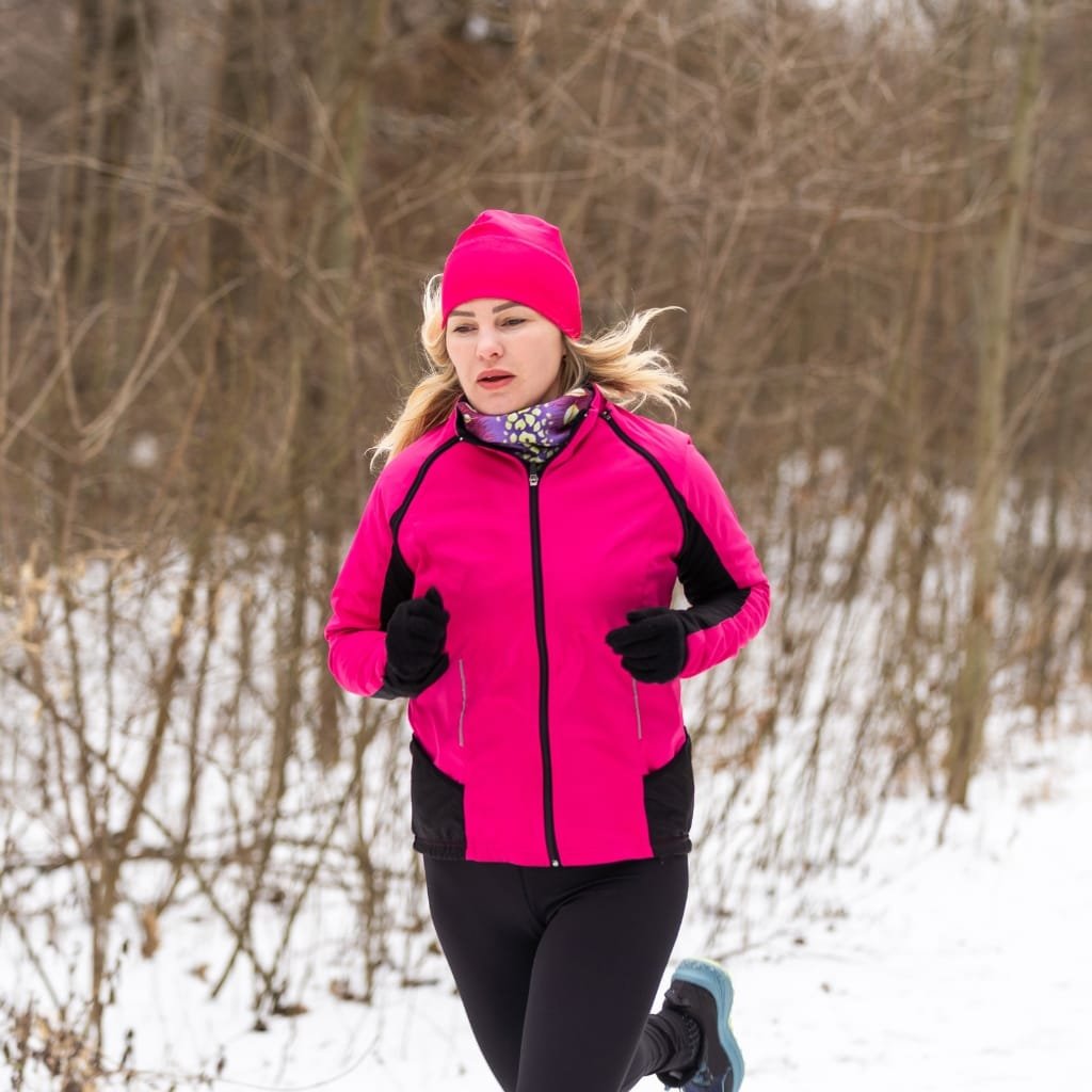 A woman running outdoors in a snowy winter landscape, dressed in a bright pink jacket, black leggings, gloves, a pink hat, and a neck warmer, with bare trees in the background.