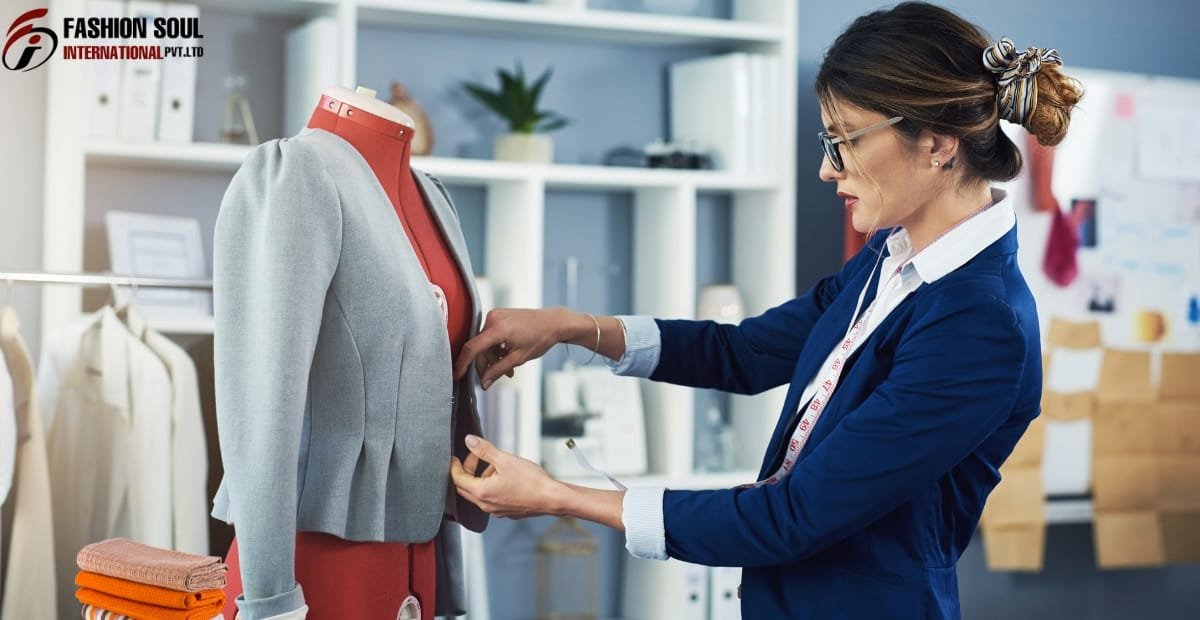 A woman in a professional setting, wearing glasses and a blue blazer, is measuring and adjusting a light gray blazer on a fashion mannequin in a design studio. The background includes shelves with fabrics, books, and design materials.
