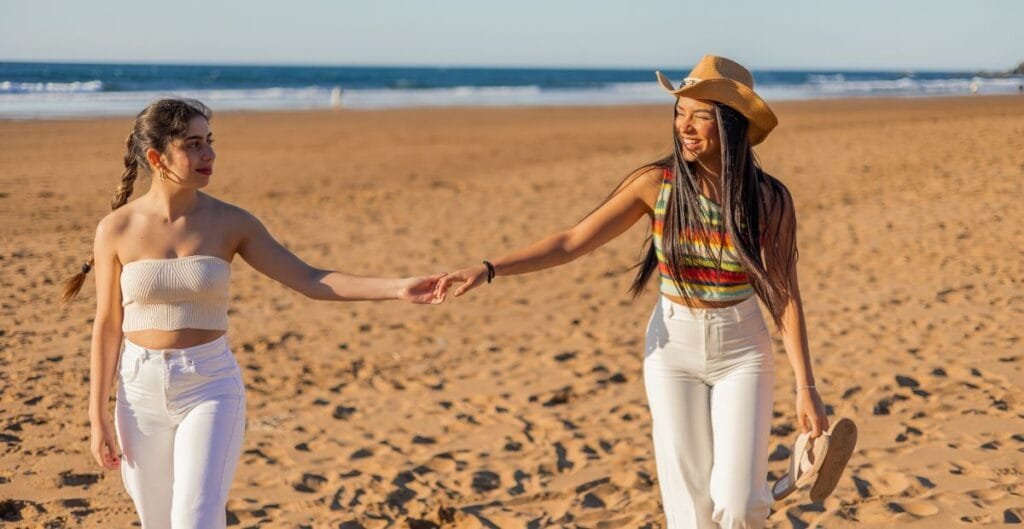 Two women walking hand-in-hand on the beach, smiling, with the ocean and clear sky in the background.