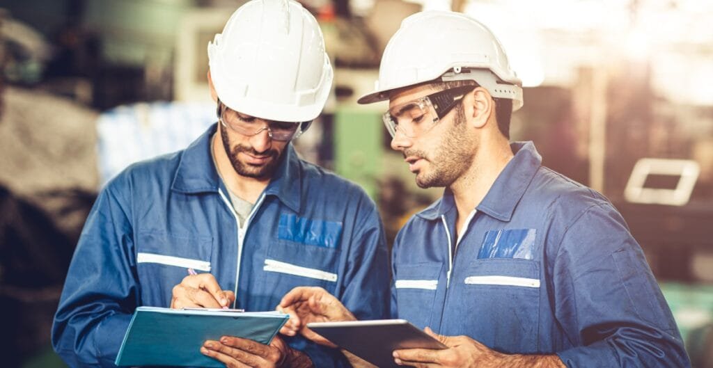 Two male industrial workers wearing safety helmets, glasses, and blue uniforms are discussing and reviewing documents in a factory setting.