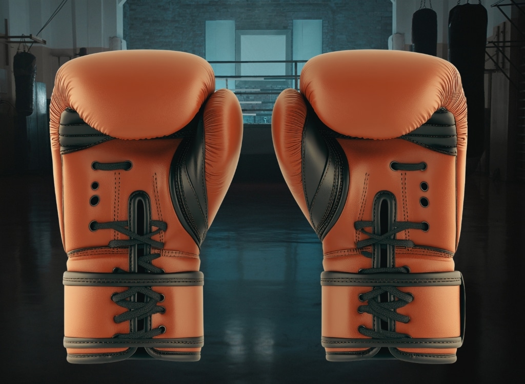 Pair of orange and black MMA gloves hanging in a gym with boxing equipment in the background.